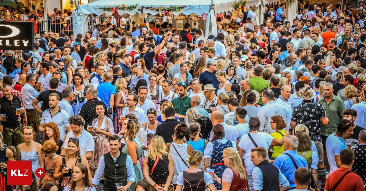 A large crowd of people gathered outdoors, possibly at a festival, with many individuals holding drinks and engaging in conversation. The setting includes tents and various stalls in the background.