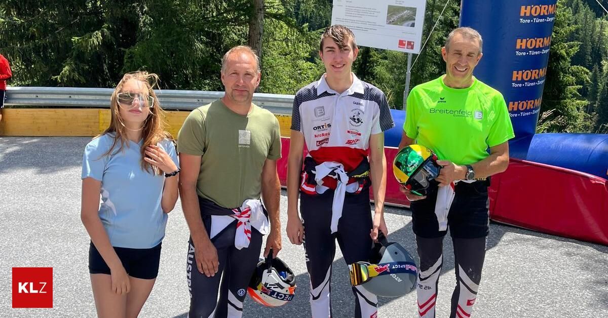 Four individuals are standing together, smiling for a photo. They are all wearing cycling gear and helmets. The man on the right is holding a helmet and a bottle. The man in the middle is holding a helmet and a piece of paper. The man on the left is holding a helmet. The woman on the left is wearing sunglasses. Behind them is a metal barrier and trees.