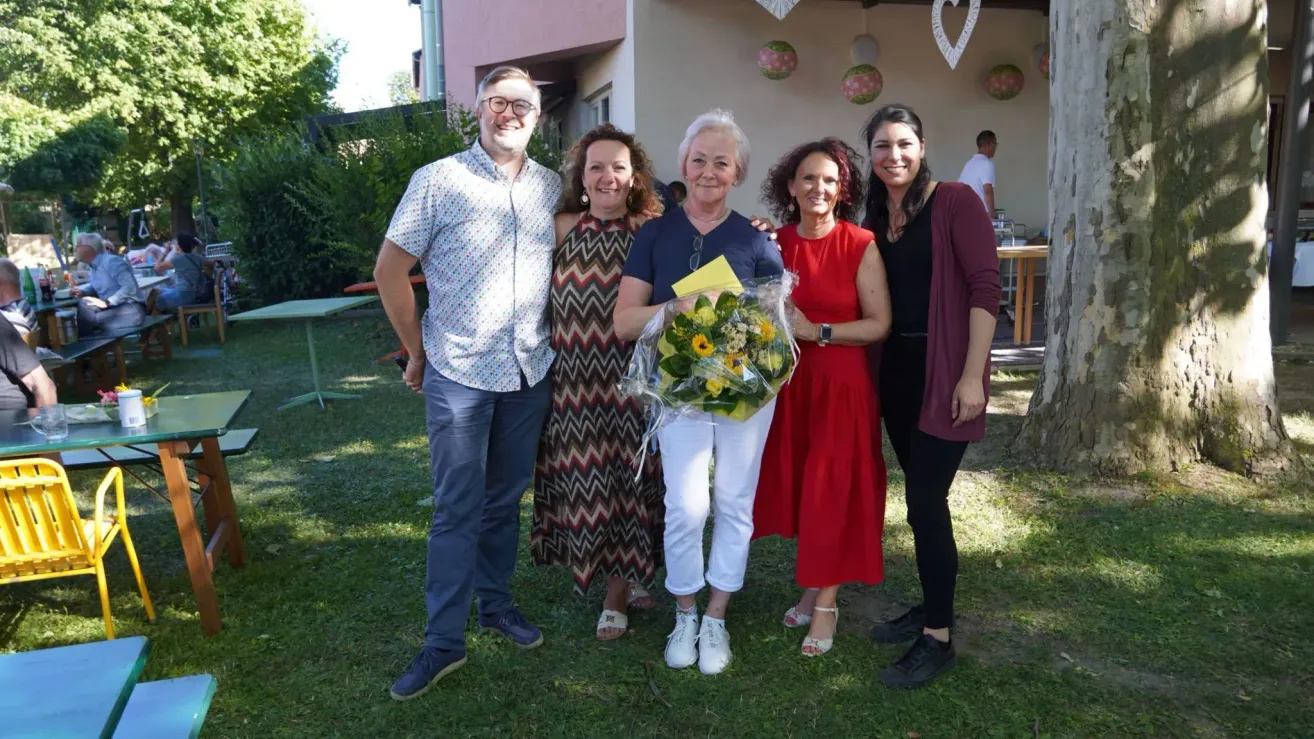 Five people are standing in a garden, smiling and posing for a photo. The woman in the middle holds a bouquet of flowers. Behind them, there is a building with decorative hangings.