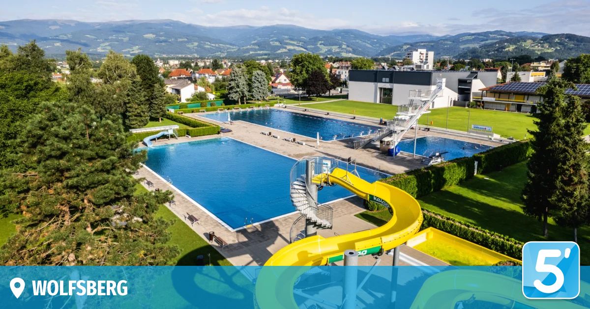 An aerial view of a large outdoor swimming pool with a yellow water slide and a diving board. The pool is surrounded by green grass and trees. In the background, there are houses and buildings, with mountains in the distance.