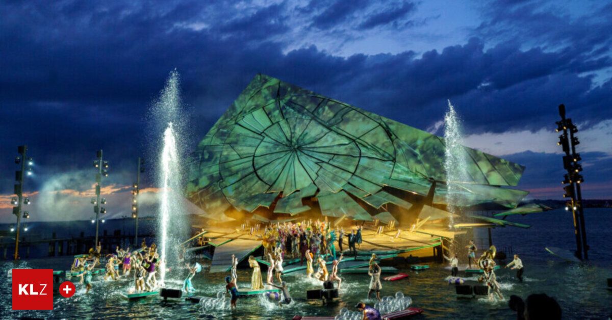 A large green structure resembling a broken shell is illuminated at night, with a crowd of people performing in the water in front of it.
