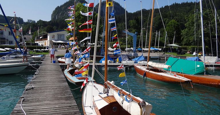 Ein Hafen mit mehreren angedockten Segelbooten, geschmückt mit Flaggen verschiedener Länder, unter einem klaren Himmel mit Bergen im Hintergrund.