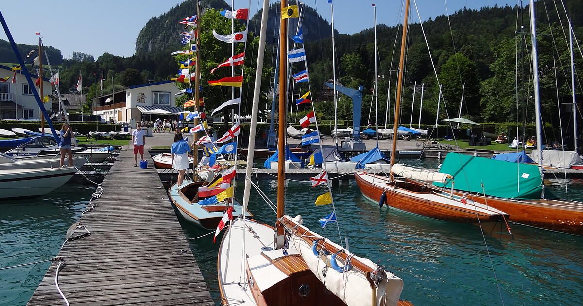 Ein Hafen mit mehreren angedockten Segelbooten, geschmückt mit Flaggen verschiedener Länder, unter einem klaren Himmel mit Bergen im Hintergrund.