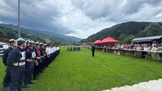 Eine Gruppe uniformierter Personen steht in einer Reihe auf einem Rasenfeld mit Bergen im Hintergrund. Zuschauer sitzen unter roten Zelten.