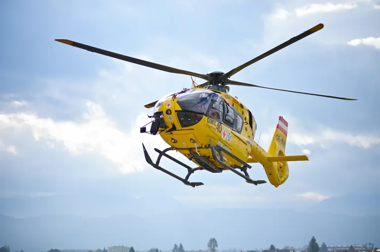 Ein gelber Hubschrauber mit einem roten Kreuz auf der Seite und der Registriernummer OE-XVI fliegt am Himmel.