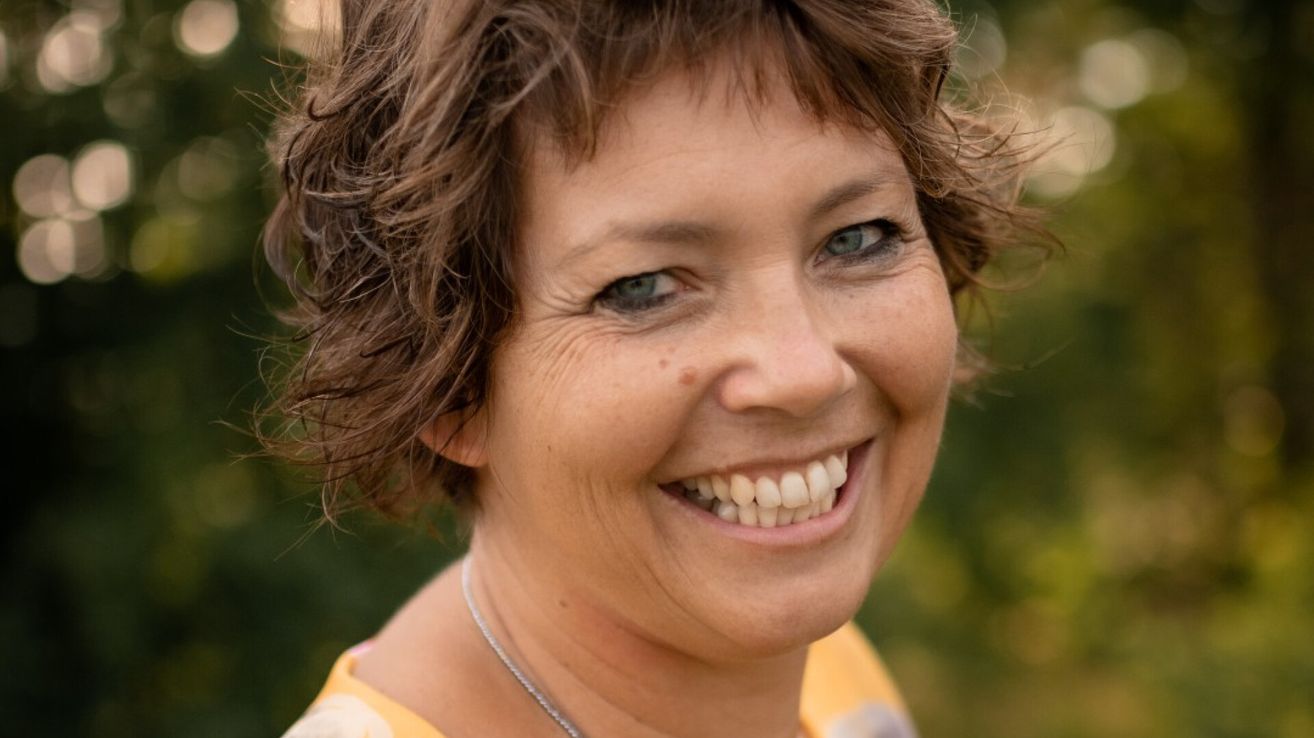 Eine Frau mit kurzen, lockigen braunen Haaren und blauen Augen lächelt fröhlich. Ihr Gesicht zeigt Altersspuren mit Falten und Sommersprossen. Sie trägt ein gelbes Oberteil und eine silberne Halskette.