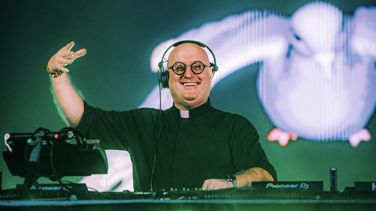 Ein lächelnder Mann mit Priesterkragen und Kopfhörern, der hinter einem DJ-Setup mit einem Pioneer-Controller steht.