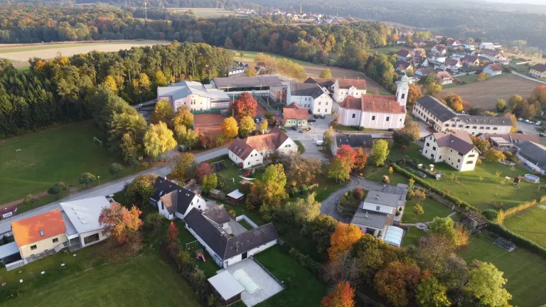 Eine Luftaufnahme eines Dorfes mit verschiedenen Gebäuden, umgeben von Bäumen und Grünflächen. Die Gebäude sind überwiegend weiß, einige haben rote Dächer. Ein Tennisplatz und ein Spielplatz sind sichtbar.
