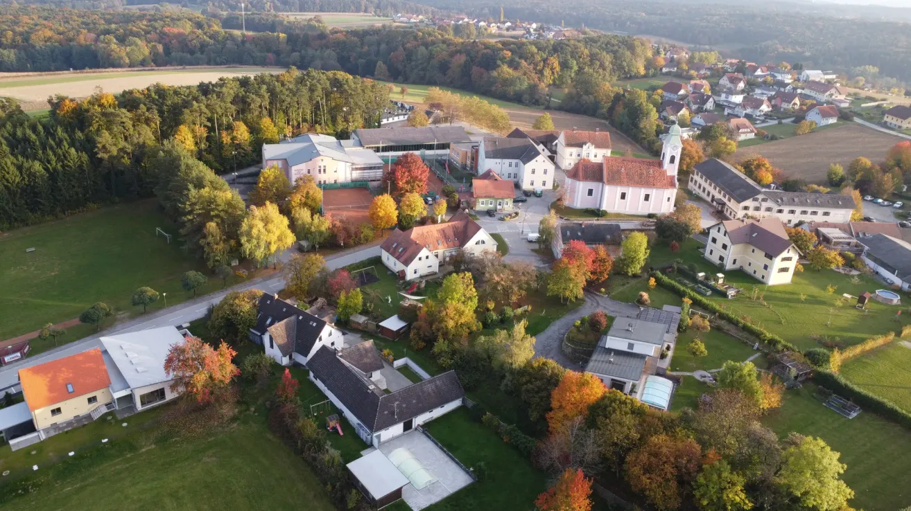 Eine Luftaufnahme eines Dorfes mit verschiedenen Gebäuden, umgeben von Bäumen und Grünflächen. Die Gebäude sind überwiegend weiß, einige haben rote Dächer. Ein Tennisplatz und ein Spielplatz sind sichtbar.