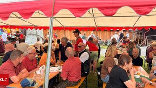 Eine Gruppe von Menschen sitzt unter einem roten Zelt um Tische herum, einige tragen Hüte und Brillen, während andere stehen und sich unterhalten. Die Umgebung scheint ein zwangloses Outdoor-Treffen zu sein, mit einer Holzstruktur und einer Kletterwand im Hintergrund.