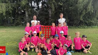 Eine Gruppe von Kindern in rosa Shirts und Mützen posiert für ein Foto auf einer Wiese mit zwei Männern hinter einem roten Spielbrett.