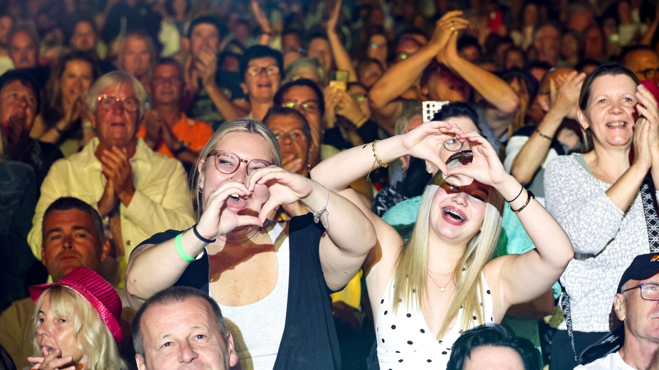 Eine Menschenmenge bei einer Veranstaltung, zwei Frauen vorne machen Herzformen mit ihren Händen, lächeln und tragen Brillen.