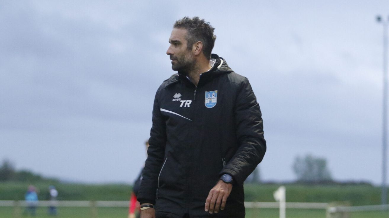 Ein Mann in einer schwarzen Jacke mit weißen und blauen Logos steht auf einem Rasenfeld und schaut nach links.