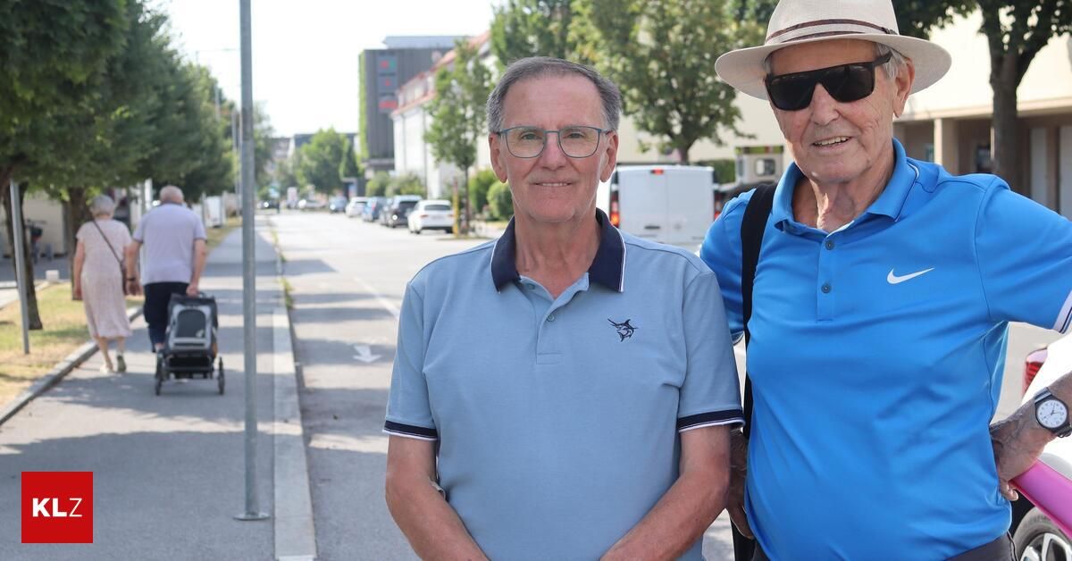 Zwei ältere Männer stehen am Straßenrand und lächeln für ein Foto. Der Mann auf der linken Seite trägt eine Brille und ein blaues Poloshirt mit Logo. Der Mann auf der rechten Seite trägt einen Hut und eine Sonnenbrille. Hinter ihnen sind geparkte Autos, ein weißer Lieferwagen und ein Gebäude.