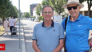 Zwei ältere Männer stehen am Straßenrand und lächeln für ein Foto. Der Mann auf der linken Seite trägt eine Brille und ein blaues Poloshirt mit Logo. Der Mann auf der rechten Seite trägt einen Hut und eine Sonnenbrille. Hinter ihnen sind geparkte Autos, ein weißer Lieferwagen und ein Gebäude.