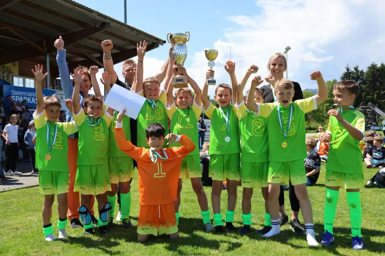 Eine Gruppe junger Fußballer in grünen Trikots mit den Nummern 1, 8 und 11 feiert mit Trophäen und Medaillen auf einem Rasenfeld.