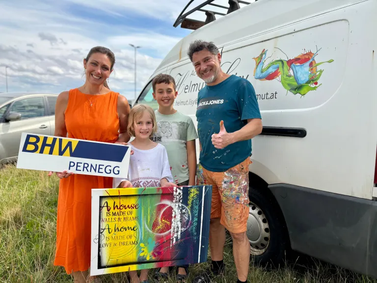 Eine vierköpfige Familie steht vor einem weißen Van und hält ein Schild mit der Aufschrift 'PERNEGG' und ein farbenfrohes Gemälde. Der Mann gibt einen Daumen hoch.
