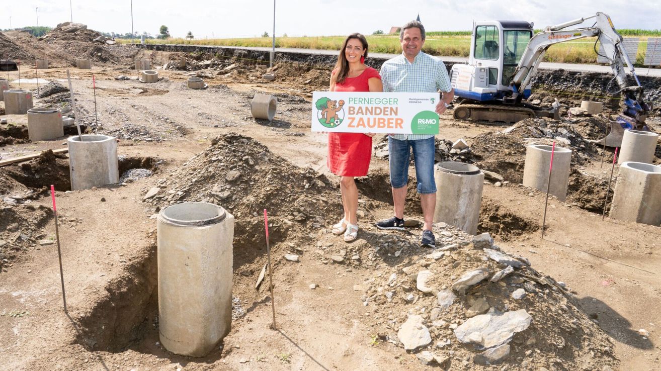 Ein Mann und eine Frau stehen auf einer Baustelle und halten ein Schild mit der Aufschrift Pernegger Banden Zauber. Dahinter steht ein großes Baufahrzeug.