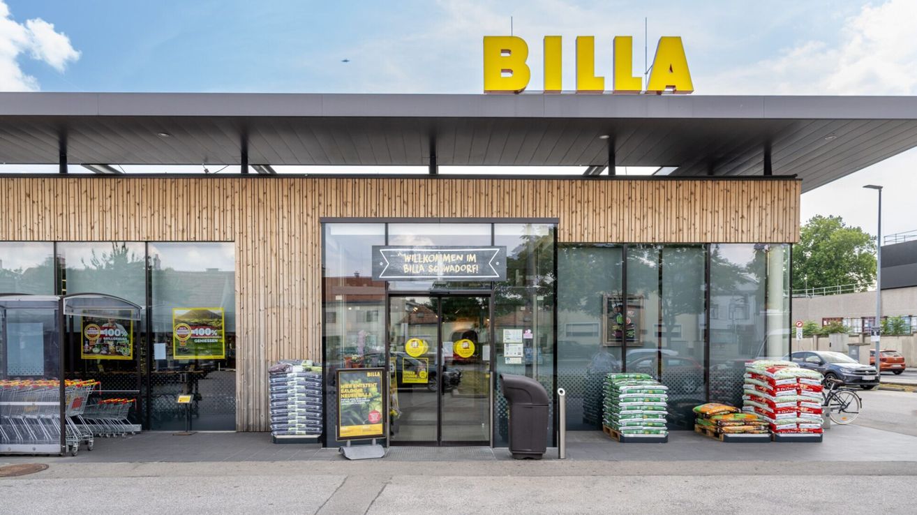Der Eingangsbereich eines BILLA-Geschäfts mit einer Glastür, einem Schild, das 'Willkommen im BILLA Schwadorf' steht, und einem Mülleimer davor.