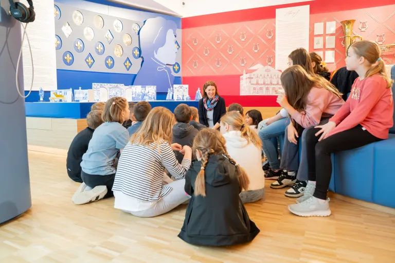 Eine Gruppe von Kindern und einer Frau sitzen auf dem Boden in einem Raum mit einer roten Wand und einer blauen Wand mit einer Silhouette einer Frau.
