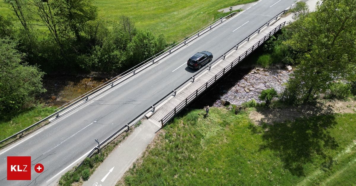 Ein schwarzes Auto fährt auf einer Straße mit einer Brücke über einen Fluss, an der Seite steht eine Person.
