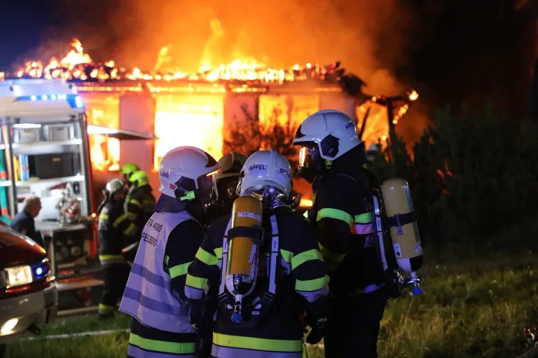 Feuerwehrleute in voller Ausrüstung stehen nachts vor einem brennenden Haus, wobei die Flammen das Gebäude verschlingen.