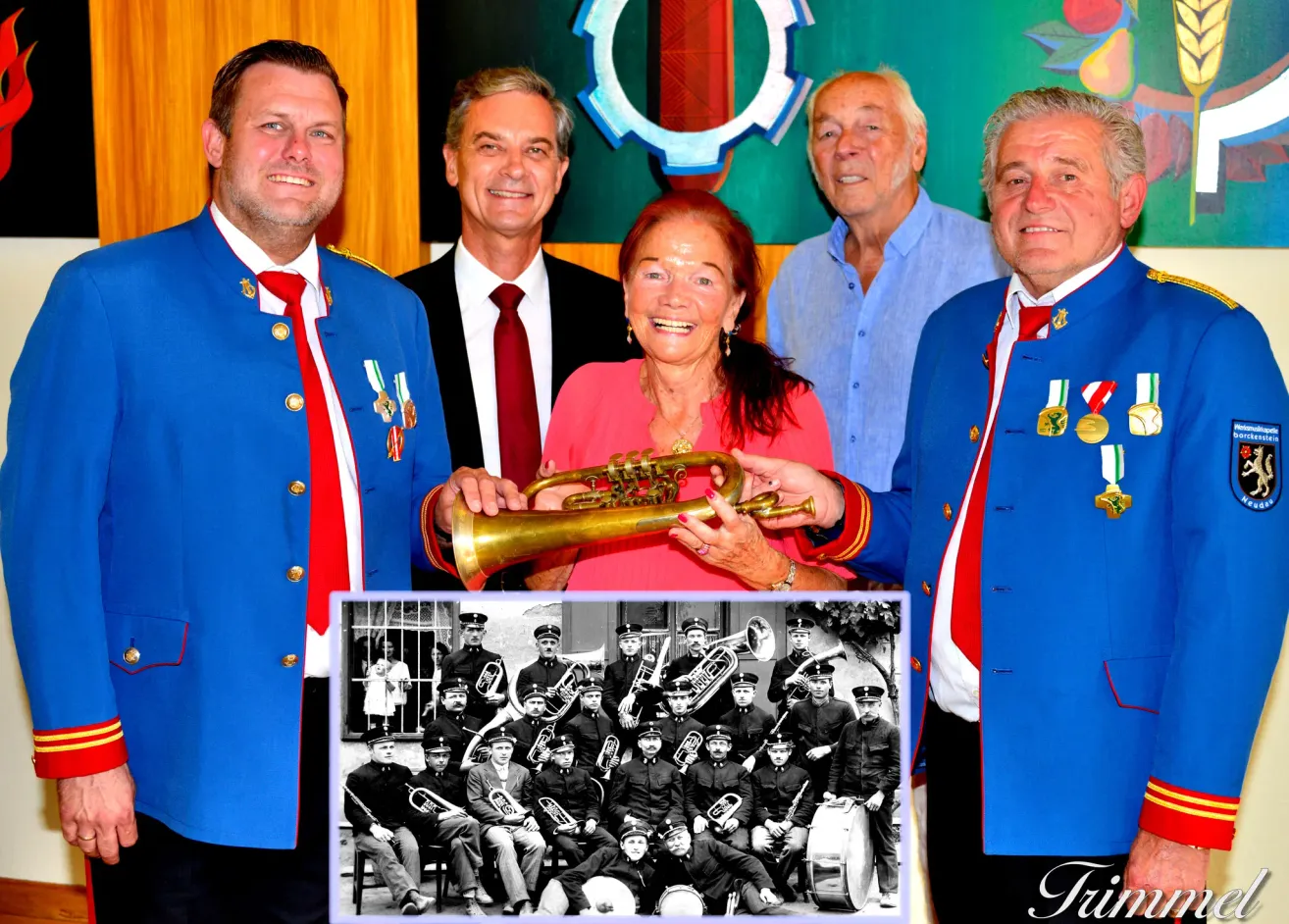 Fünf Männer und eine Frau in Uniformen und mit Medaillen, lächelnd und halten eine Trompete. Ein Schwarz-Weiß-Foto einer Marschkapelle ist eingefügt.
