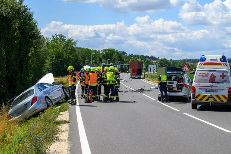 Rettungskräfte in Warnwesten stehen neben einem demoliert Auto am Straßenrand, ein LKW und ein Polizeiwagen sind in der Nähe.