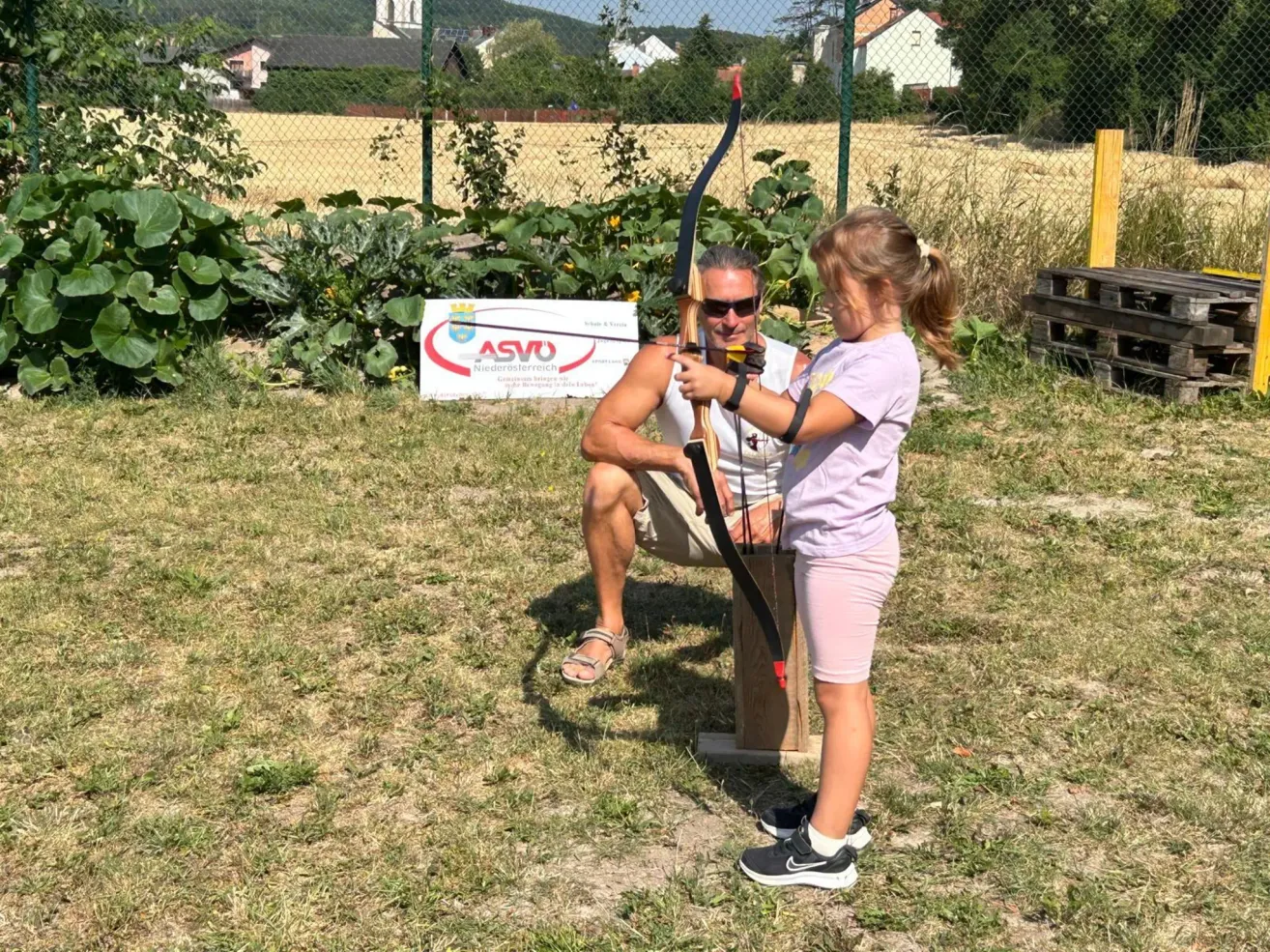 Ein Mann und ein junges Mädchen üben Bogenschießen in einem Außenbereich. Der Mann kniet auf einer Holzplattform, während das Mädchen neben ihm steht und einen Bogen hält.