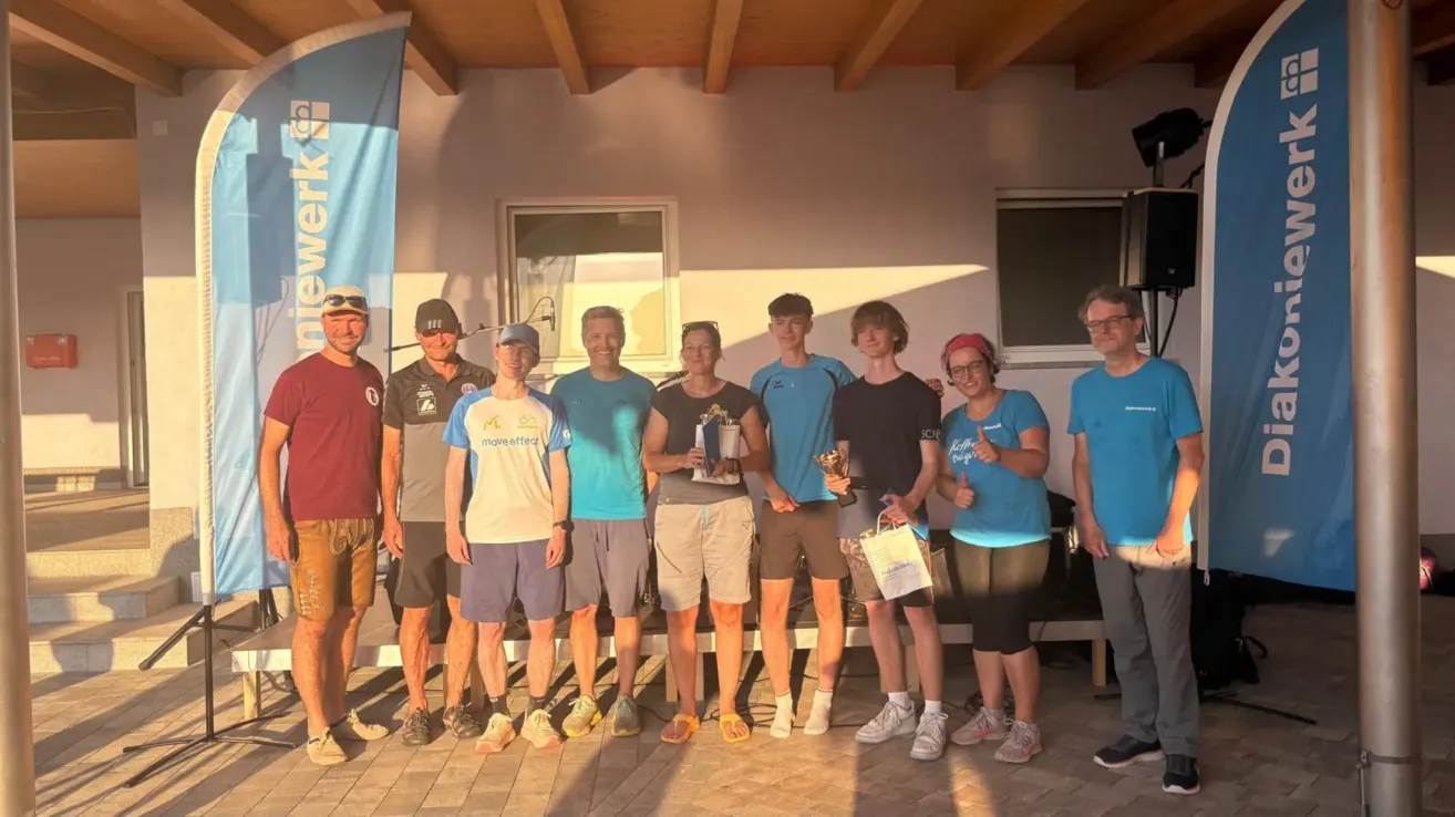 Eine Gruppe von Menschen, möglicherweise Athleten, steht zusammen und posiert für ein Foto. Sie halten Trophäen und Taschen. Sie tragen Freizeitkleidung, und einige von ihnen tragen Hüte. Hinter ihnen ist eine Wand mit einem Fenster und einem Banner.