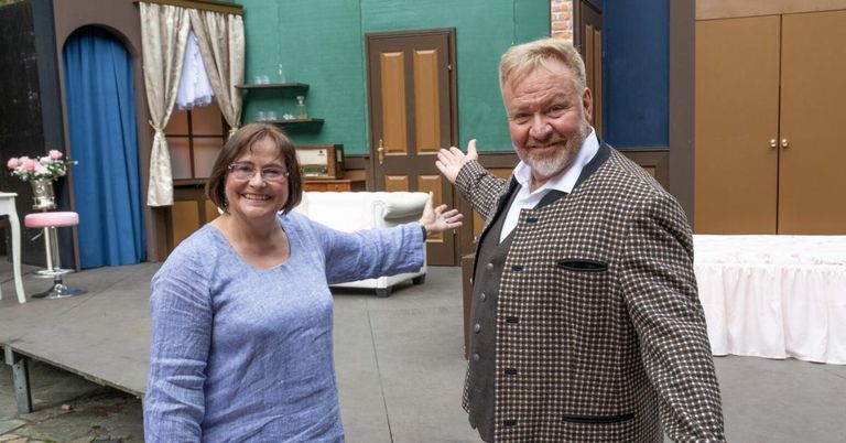 Ein Mann und eine Frau stehen in einem Raum mit einem weißen Sofa, lächeln und winken. Der Mann trägt eine karierte Jacke und die Frau ein blaues Hemd. Hinter ihnen befindet sich eine grüne Wand mit einem Regal und einem Fenster.