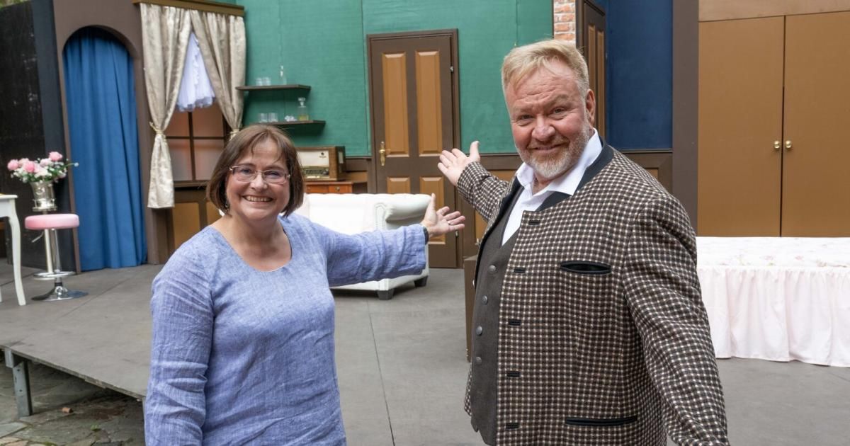 Ein Mann und eine Frau stehen in einem Raum mit einem weißen Sofa, lächeln und winken. Der Mann trägt eine karierte Jacke und die Frau ein blaues Hemd. Hinter ihnen befindet sich eine grüne Wand mit einem Regal und einem Fenster.