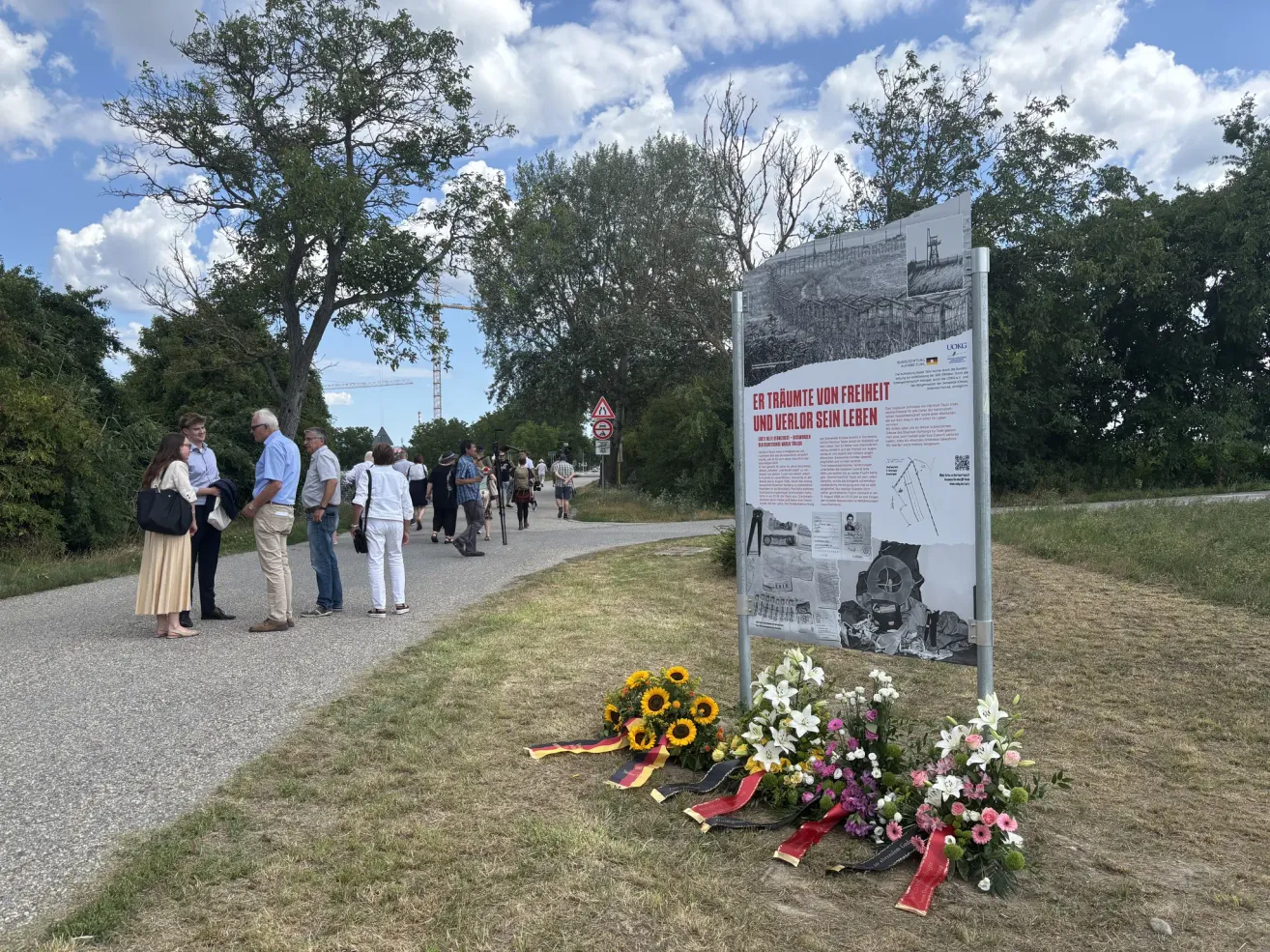 Ein Gedenkschild steht auf einem Grasbereich mit Blumen und Bändern. Mehrere Menschen stehen und gehen auf einem Pfad in der Nähe. Das Schild hat Text auf Deutsch.