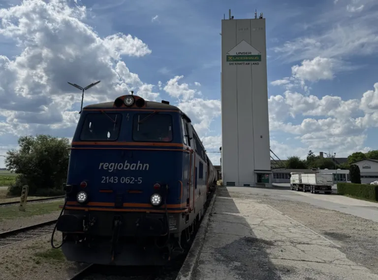 Ein blauer Regiobahn-Zug steht auf den Gleisen, mit einem hohen Lagerhaus-Silo im Hintergrund.