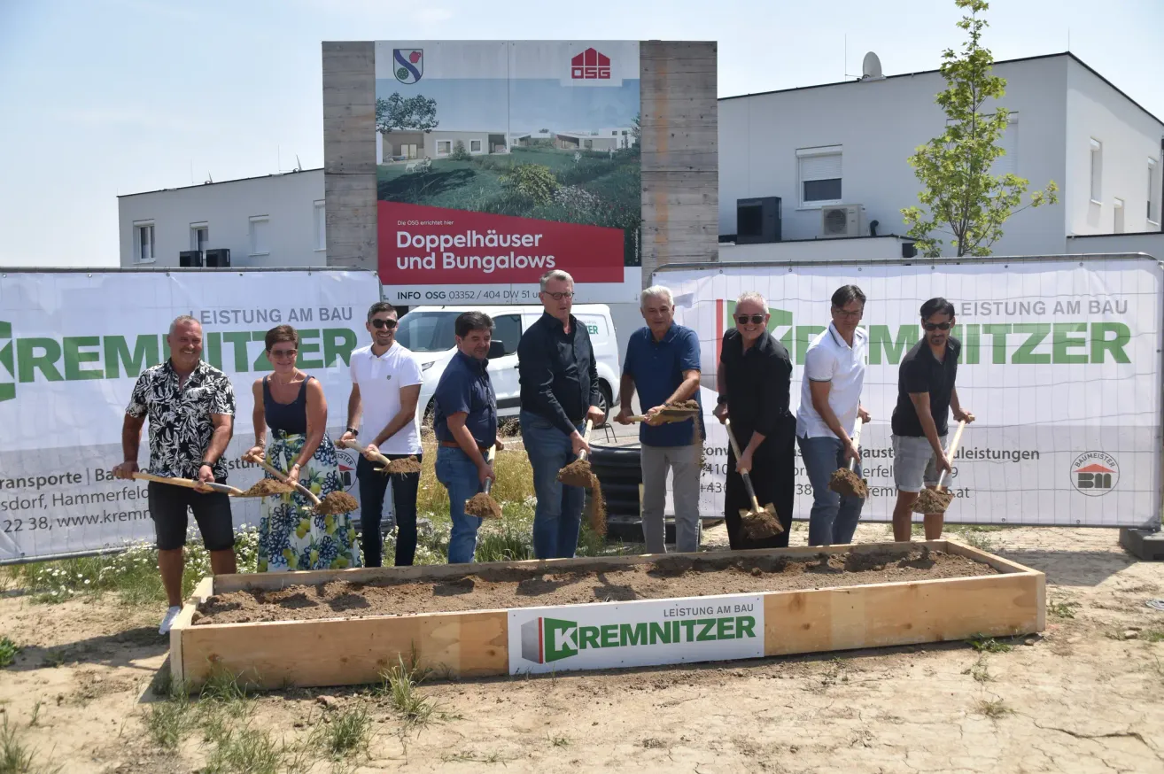 Eine Gruppe von Menschen in formeller Kleidung steht in einer Erdgrube, hält Schaufeln und posiert für ein Foto. Im Hintergrund ist ein großes Schild mit der Aufschrift 'Doppelhauser und Bungalows' zu sehen.