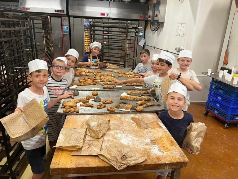 Eine Gruppe von Kindern in Kochmützen und Schürzen steht um einen Tisch mit verschiedenen Gebäckstücken und Brot in einer Küche.