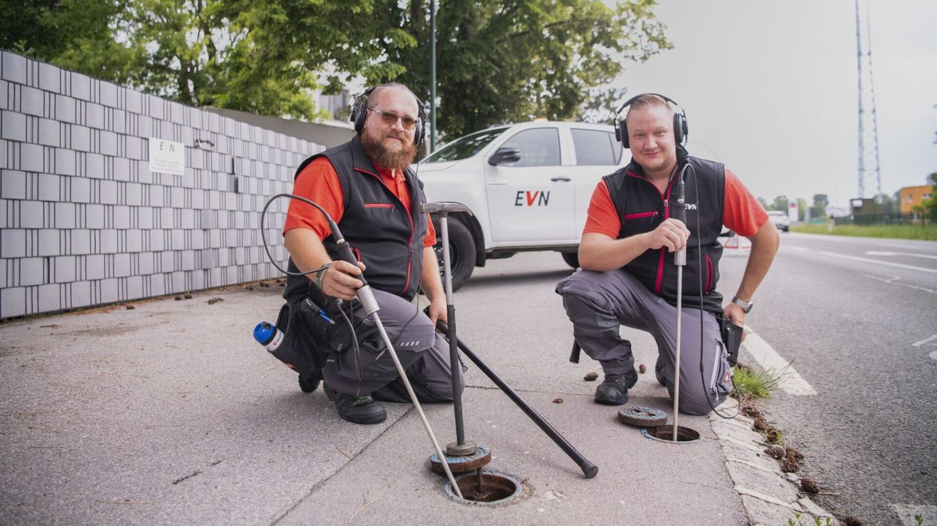 Zwei Männer in Warnwesten und Kopfhörern knien auf dem Boden, einer hält ein Werkzeug über einem offenen Kanaldeckel. Ein weißer Lastwagen mit den Buchstaben EVN ist dahinter geparkt.