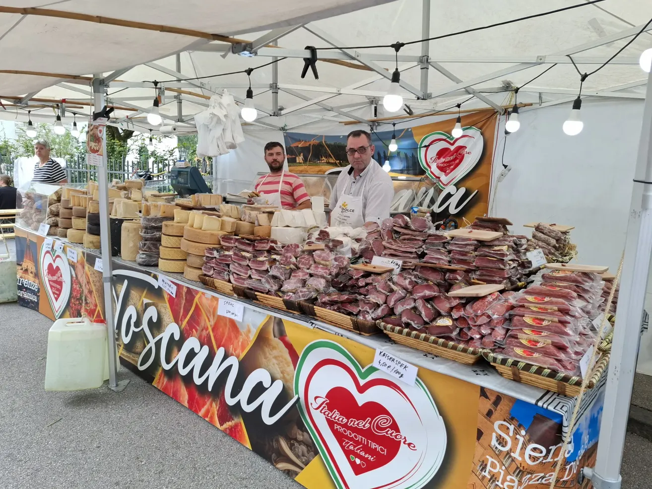 Zwei Männer stehen hinter einem Tisch voller verschiedener Fleisch- und Käsesorten, unter einem Zelt mit einem Banner, das 'Toscana' liest.