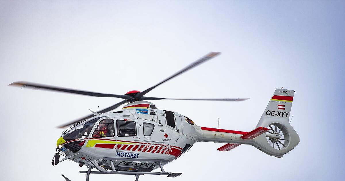 Ein weißer und roter Hubschrauber mit der Registrierung OE-XW ist im Flug mit einem Piloten im Inneren. Der Hubschrauber hat ein rotes Kreuzsymbol an der Seite.