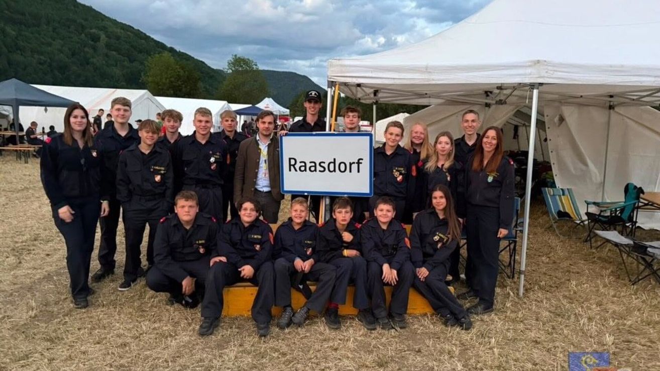Eine Gruppe uniformierter Personen, einschließlich Erwachsener und Kinder, posiert für ein Foto vor einem Schild, das 'Raasddorf' steht. Sie stehen und sitzen auf einer gelben Plattform, mit Zelten und einem Berg im Hintergrund.
