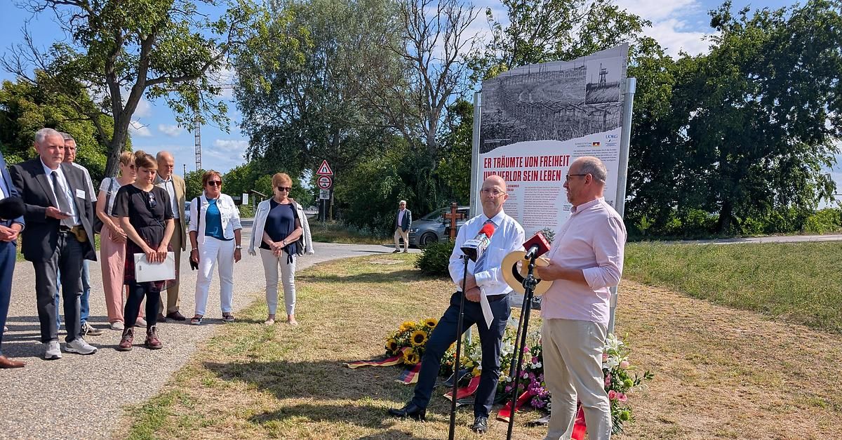 Eine Gruppe von Menschen steht um ein Denkmal, zwei Männer mit Mikrofonen sprechen, während andere zuhören. Ein großes Schild mit deutschem Text steht dahinter.