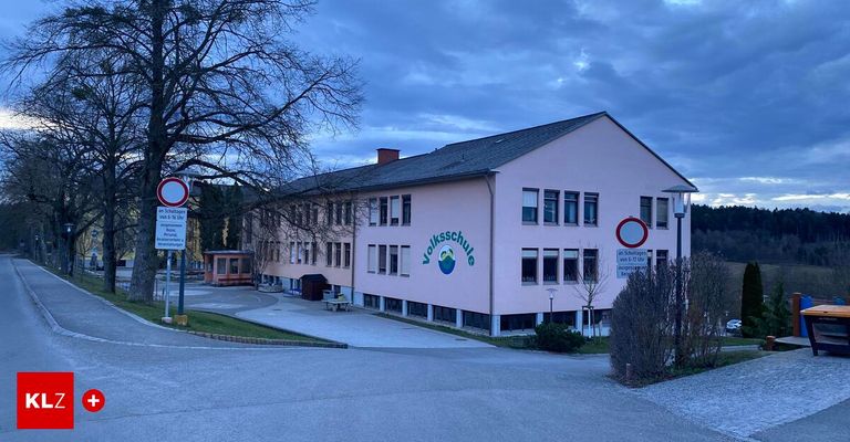 Ein rosafarbenes Gebäude mit dem Schild 'Volksschule' an der Wand, einer Bank davor und einem bewölkten Himmel im Hintergrund.