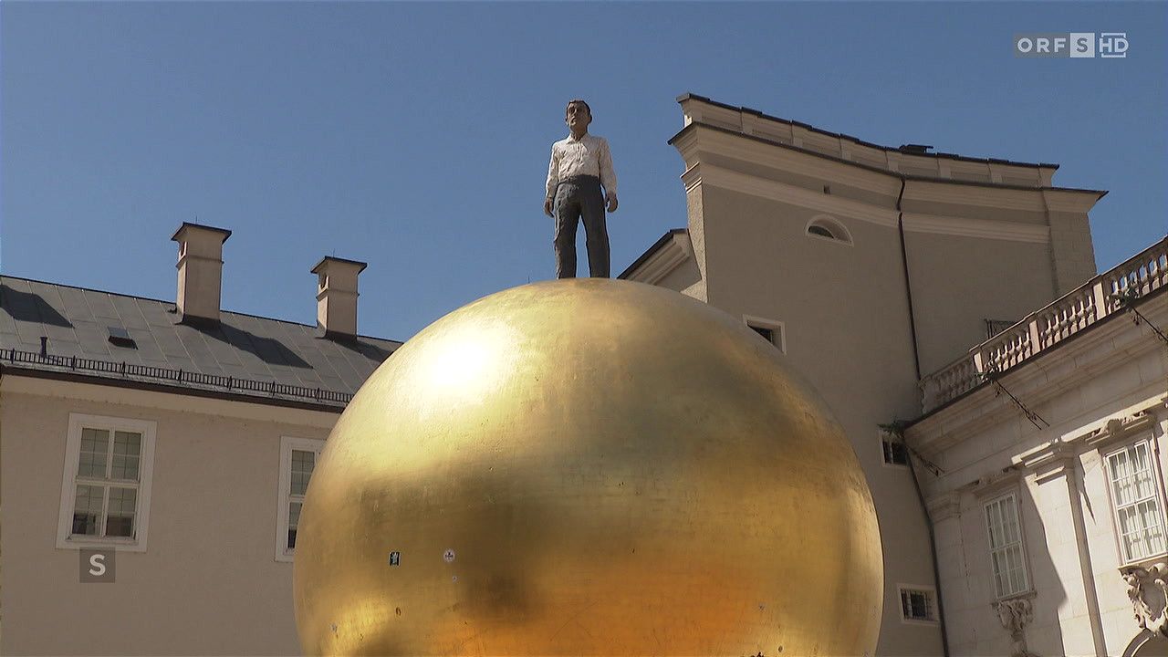 Eine goldene Kugelskulptur mit einem Mann obenauf, gekleidet in einem weißen Hemd und Hose, vor einem Gebäude mit einem Schornstein.
