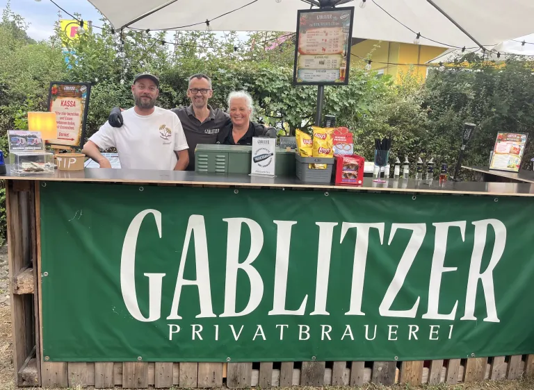 Drei Personen stehen hinter einem Tresen an einem Bierstand mit einem Banner, das 'GABLITZE PRIVATBRAUEREI' liest. Der Tresen hat Snacks, Getränke und ein Menü.