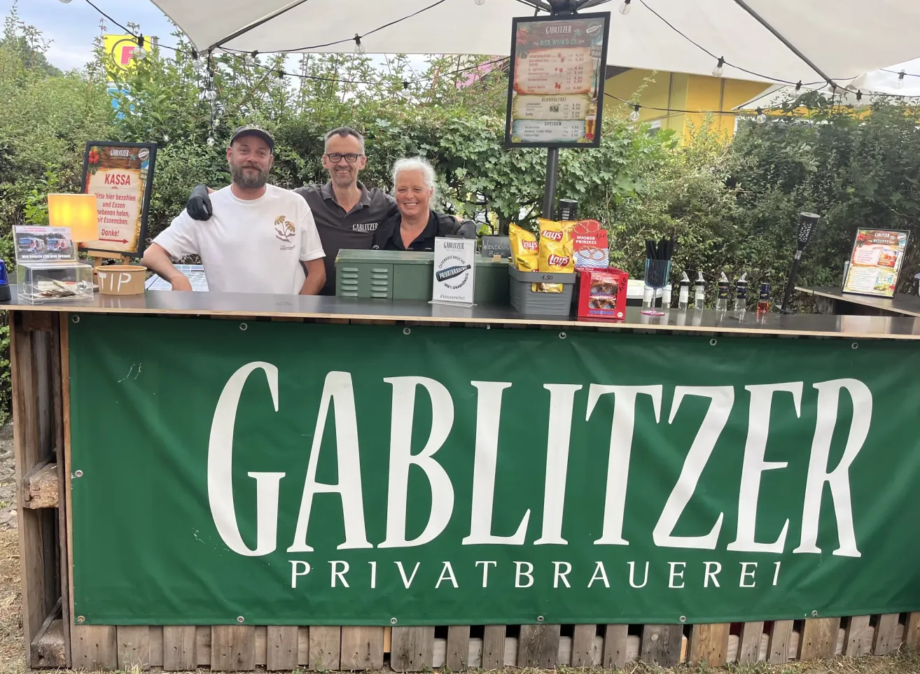 Drei Personen stehen hinter einem Tresen an einem Bierstand mit einem Banner, das 'GABLITZE PRIVATBRAUEREI' liest. Der Tresen hat Snacks, Getränke und ein Menü.