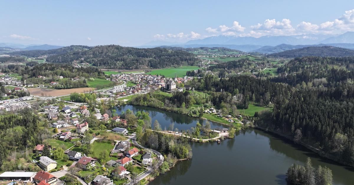 Luftaufnahme eines malerischen Dorfes zwischen einem Fluss und üppigen grünen Hügeln, mit einem Schloss in der Ferne sichtbar.