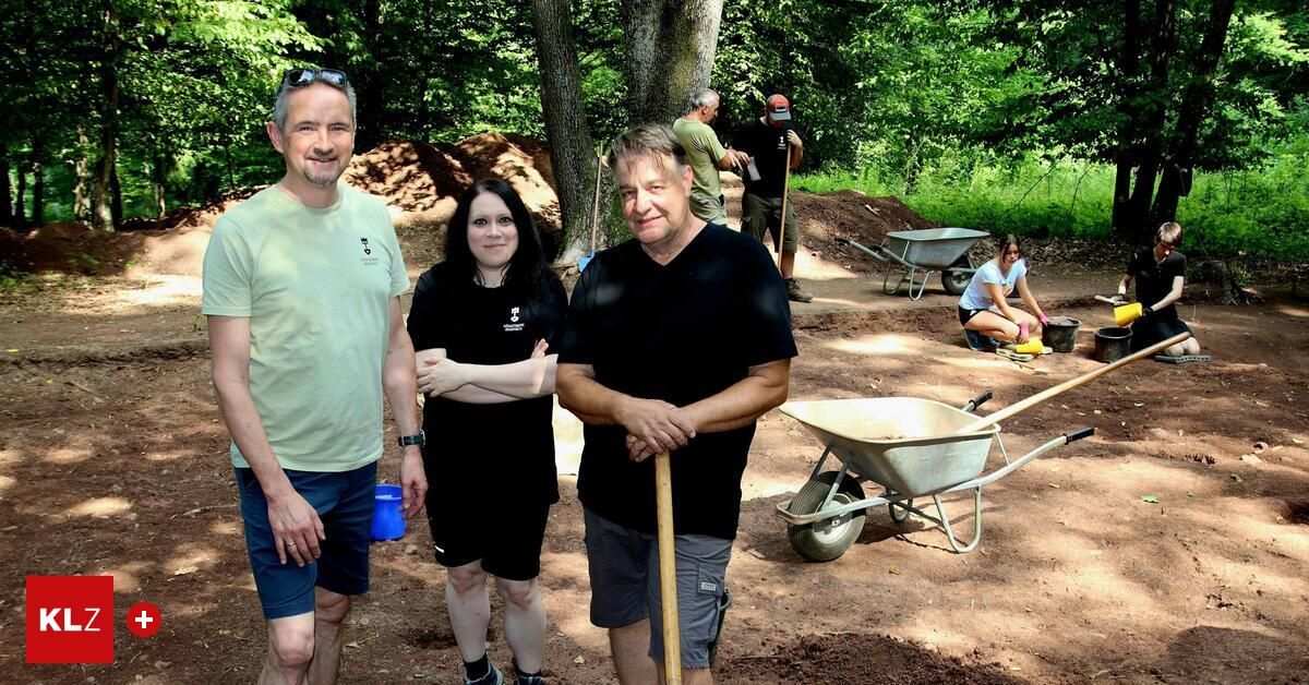 Drei Personen stehen in einem staubigen Bereich mit einem Schubkarren und einer Schaufel. Sie lächeln und posieren für ein Foto. Hinter ihnen arbeiten andere Personen auf dem Boden. Es gibt Bäume und Pflanzen in der Umgebung.