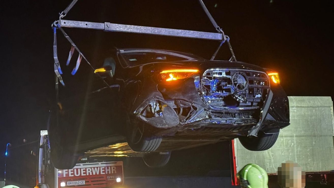 Ein schwer beschädigtes Auto wird von einem Kran angehoben und zeigt erhebliche Frontschäden mit freiliegenden Scheinwerfern und Stoßstange. Das Fahrzeug befindet sich auf einem Flachbett-Lkw mit einem roten Schild, das 'FEUERWEHR' besagt.