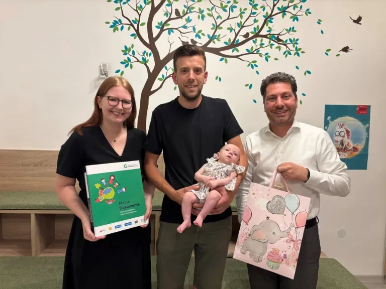 Eine dreiköpfige Familie steht vor einer Wand mit einem Baum-Wandbild und hält ein Baby, ein Buch und einen Geschenkbeutel.