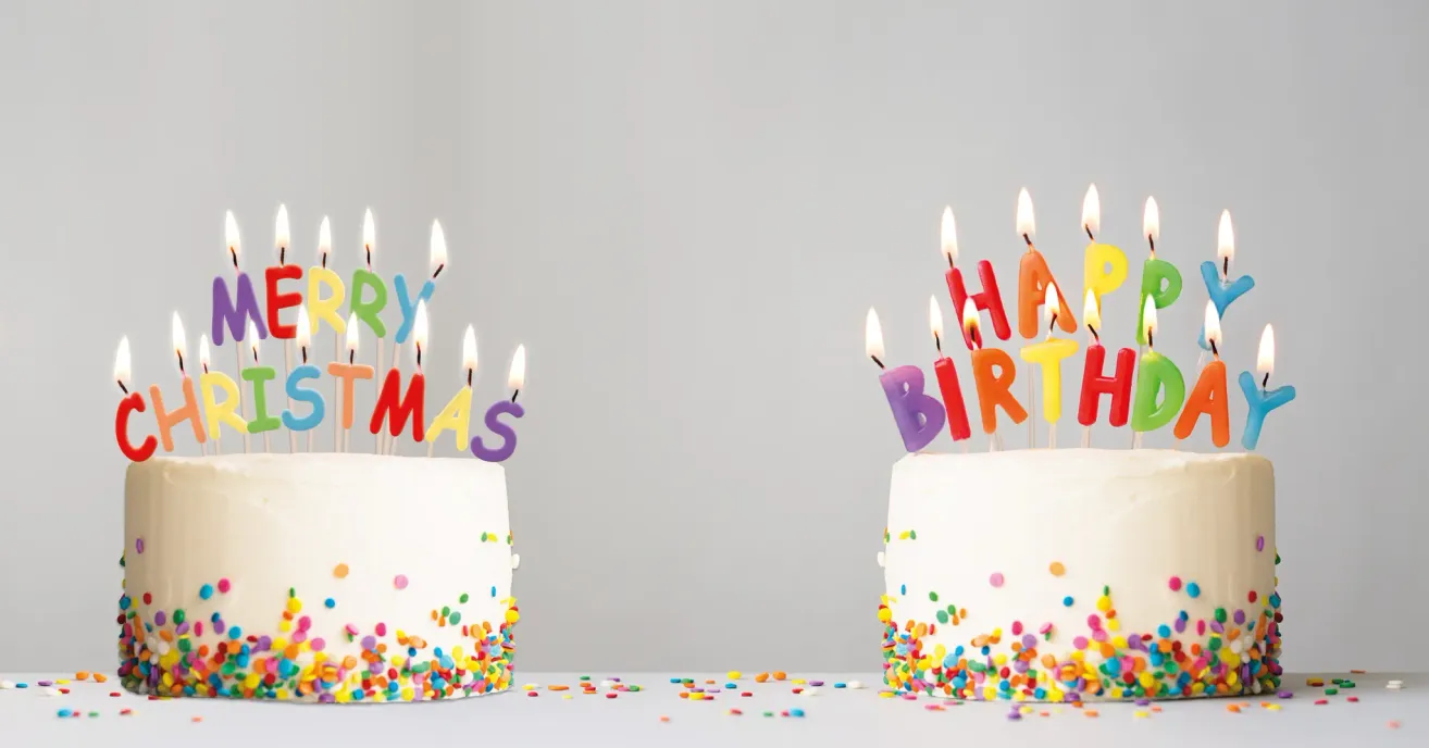 Zwei Geburtstagskuchen mit bunten Streuseln und Kerzen, einer mit 'Frohe Weihnachten' und der andere mit 'Alles Gute zum Geburtstag' obenauf geschrieben.